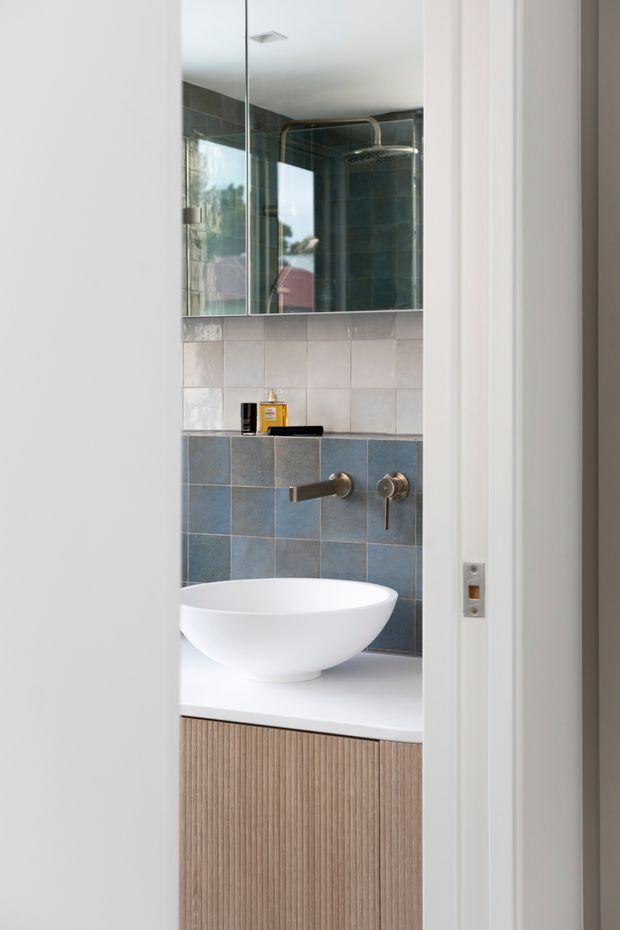 A compact contemporary bathroom viewed through a partially open doorway. The room features a white vessel basin on a fluted timber vanity, blue-grey and pale square wall tiles, a wall-mounted bronze-toned tap, mirrored upper wall, and a ceiling-mounted shower visible in the reflection. Natural light enters through a window reflected in the mirror.