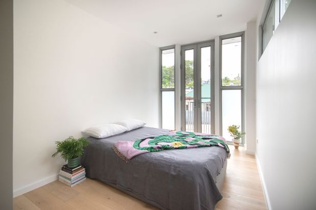 A bright, minimalist bedroom with a low-profile bed dressed in dark grey bedding and a colourful patterned throw. Floor-to-ceiling windows and glazed doors provide abundant natural light and views toward neighbouring buildings. Light timber flooring, white walls, simple recessed ceiling lights, potted plants, and stacked books create a clean, relaxed interior.