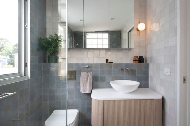 Contemporary bathroom with a wall-mounted vanity, white vessel basin, concealed-cistern toilet, frameless glass shower screen, large mirrored cabinet, and round wall sconce. The space uses pale grey-blue square wall tiles, white fixtures, and a fluted light-wood vanity front, with natural light entering through a side window.