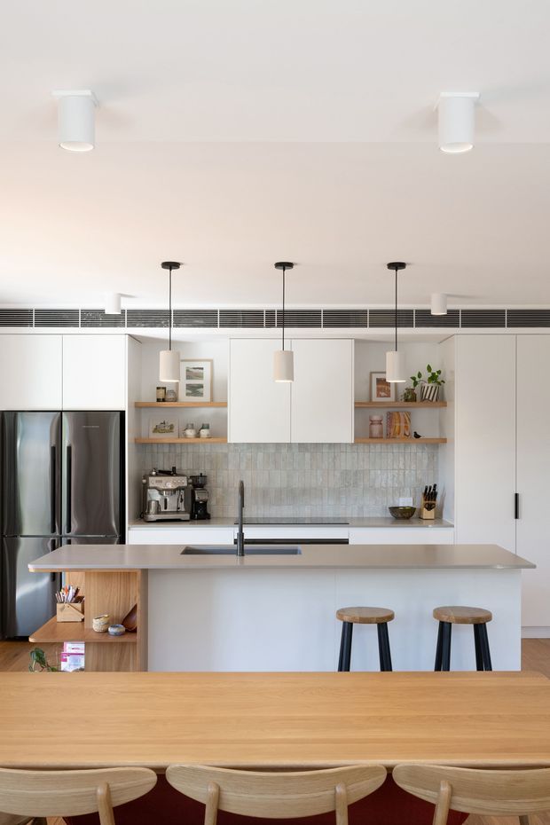 Bright contemporary kitchen with a large central island, integrated stainless-steel refrigerator, white flat-panel cabinetry, open timber shelves, pale vertically textured tile splashback, and three pendant lights. A timber dining table with curved wooden chairs occupies the foreground. The room has a clean, warm modern fit-out with concealed linear air-conditioning grilles and recessed ceiling spotlights.