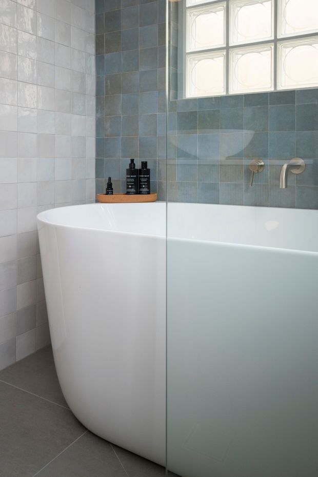 A bright, contemporary bathroom featuring a freestanding white bathtub enclosed by a clear glass shower screen. The walls are clad in pale grey and muted blue-grey square tiles, with dark grey floor tiles. A small frosted decorative window provides natural light, and brushed metal wall-mounted tapware is visible above the tub.