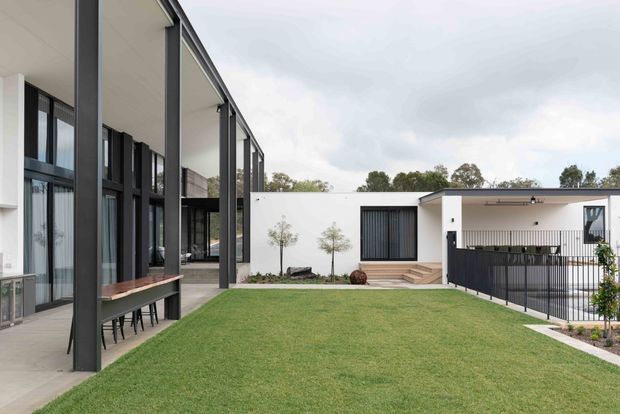 A contemporary residential courtyard viewed from a covered outdoor area, with a broad lawn enclosed by low white rendered building wings. Tall dark structural posts and full-height glazing line the left side, while the right wing has a flat roof, dark-framed windows, a small terrace, and black metal balustrading. The scene has clean geometric lines and a restrained monochrome palette with green landscaping.