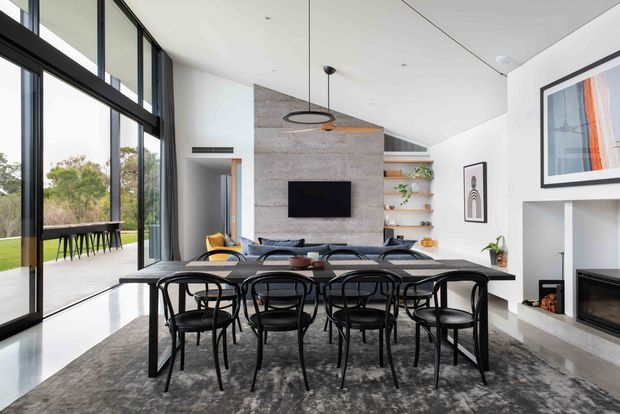 Bright contemporary open-plan dining and living area with a large black-framed glazed wall opening to the garden. A long dark dining table with multiple black chairs occupies the foreground, while a lounge, wall-mounted television, built-in shelving, fireplace and concrete-look feature wall sit beyond. The space has a high ceiling, polished light floor, and restrained black, white, grey and timber palette.
