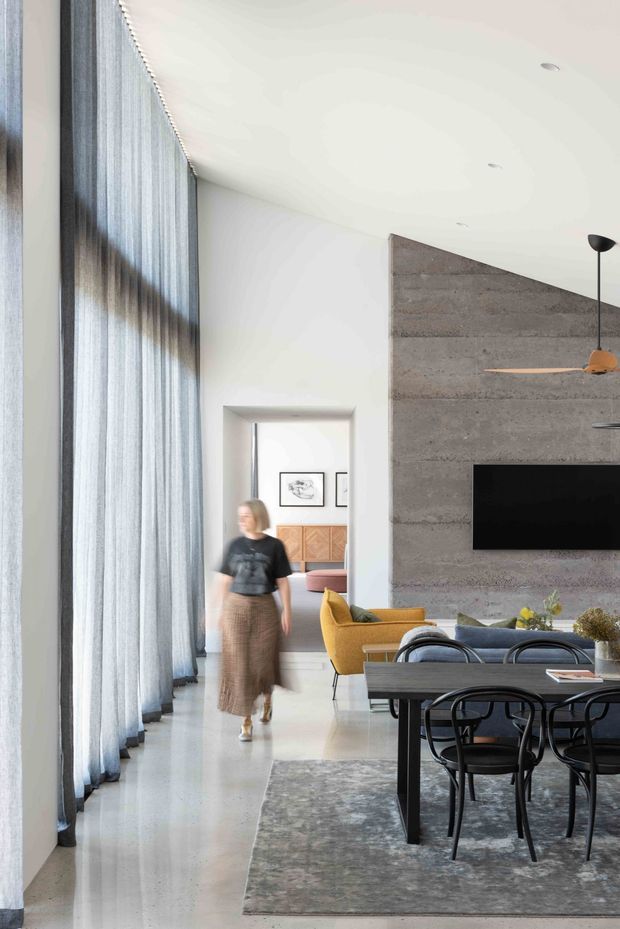 A bright, spacious open-plan contemporary interior combining a dining area and living room. Full-height sheer curtains line the left side, while a textured grey concrete feature wall with a wall-mounted television anchors the living zone. A dark dining table with black chairs sits on a large grey rug, alongside mustard-yellow and blue-grey upholstered seating. White walls and polished pale flooring create an airy, refined atmosphere, with a glimpse of an adjoining furnished room beyond.