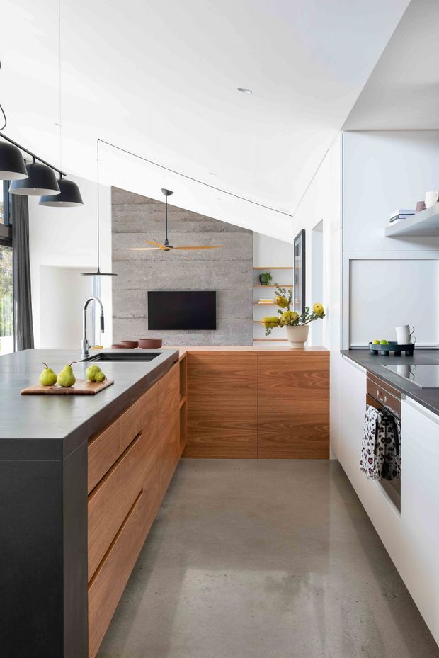 Bright contemporary kitchen with a long central island, warm timber cabinetry, dark benchtops, white walls and integrated appliances. A grey concrete feature wall with a wall-mounted television forms the backdrop, while pendant lights, open shelving and a sculptural ceiling add visual interest.