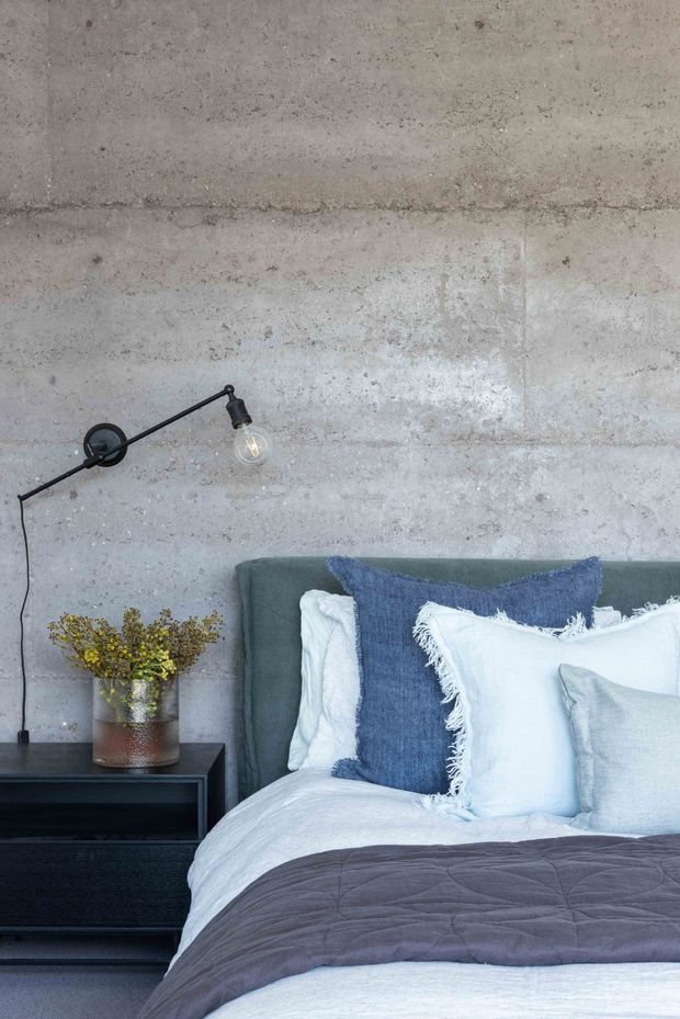 Close-up interior view of a contemporary bedroom, featuring a partially visible upholstered bed with layered white, blue, and grey bedding, a dark bedside table, and a slim black wall-mounted reading light with an exposed bulb. The backdrop is a raw, mottled grey concrete wall, creating an industrial bedroom aesthetic.