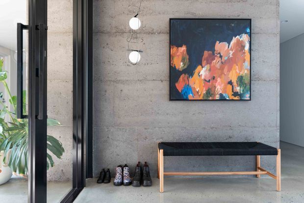 A contemporary entryway with a textured exposed concrete feature wall, black-framed glazed door, hanging globe pendant lights, a timber-framed upholstered bench, footwear storage, and colourful abstract artwork. A leafy indoor plant is visible through the glazing.