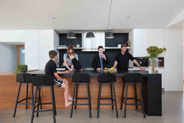 A bright, contemporary open-plan kitchen centred on a large timber island with dark cabinetry and multiple black bar stools. The kitchen includes a dark stone-look benchtop, white full-height cabinetry, a black tiled or painted feature backsplash, integrated appliances, and three pendant lights. Several people gather around the island, with a dining area and doorway visible beyond.