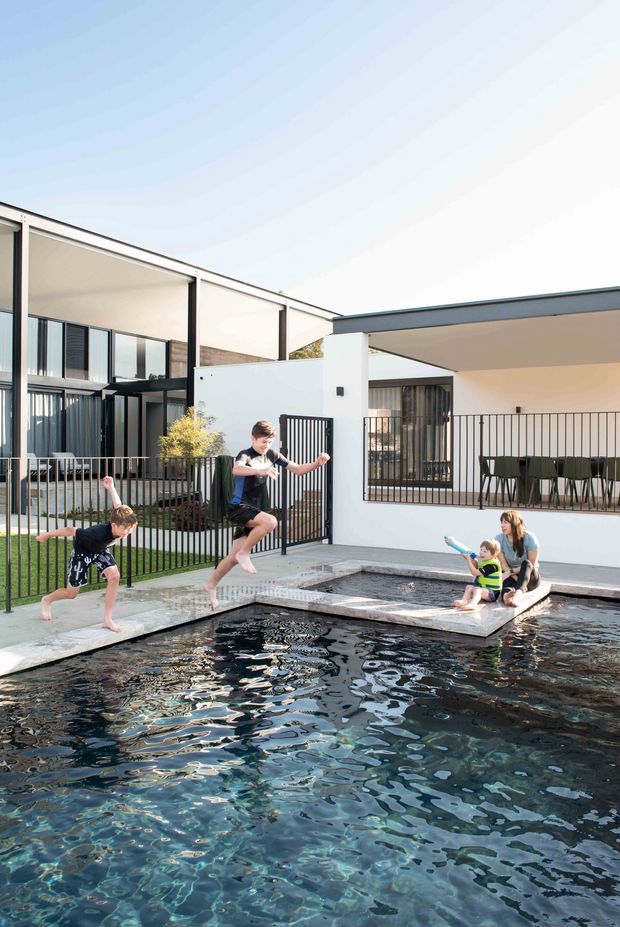 Bright exterior view of a contemporary multi-level residence centred around a large swimming pool and smaller adjoining pool or spa. The home has crisp white rendered walls, dark metal window and door frames, open covered terraces, black metal balustrades, and a flat roofline. Children and adults are using the pool area, which is bordered by concrete paving and landscaped planting.