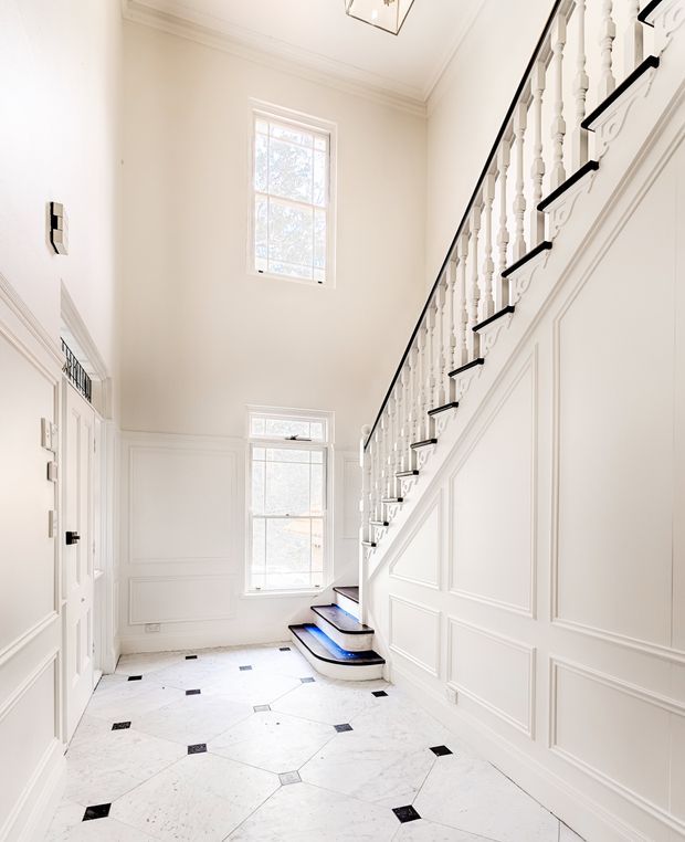 Bright, elegant stairway and entry hall with a sweeping staircase, white painted turned balusters, black stair nosings, and extensive white decorative wall panelling. The floor is a light marble or stone surface with small black diamond insets. Tall windows bring in abundant natural light, while classic moulded cornices and trim reinforce the refined traditional character.