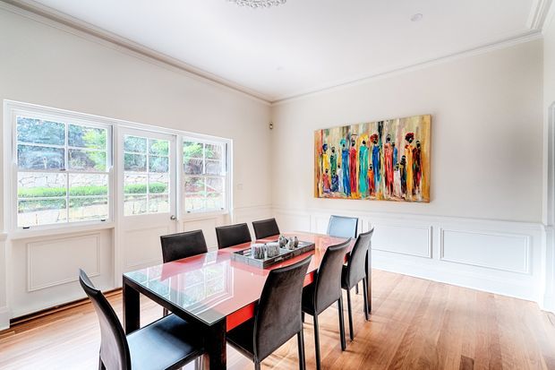 Bright, elegant dining room with a long glass-topped dining table and black upholstered chairs. The room features pale neutral walls with decorative white wainscoting and crown moulding, tall multi-pane windows and glazed doors, polished timber flooring, and a colourful abstract artwork as the main accent.