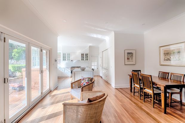 Bright open-plan interior combining a dining area, living seating and a white kitchen. The room features warm timber flooring, white walls and cabinetry, large glazed French doors opening to an outdoor terrace, a dark timber dining table with matching chairs, woven armchairs, and framed artwork. The space has a refined traditional character with abundant natural light.