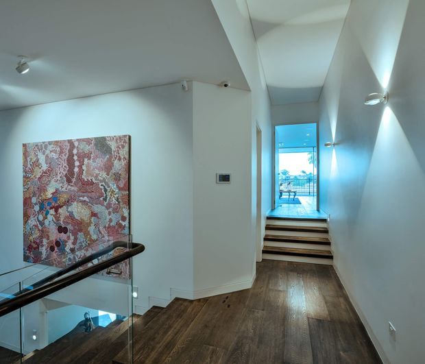 Interior view of a contemporary hallway and stair landing with dark timber flooring, white walls, recessed and wall-mounted lighting, and a glass balustrade overlooking the stairwell. A large colourful artwork forms a feature on the left wall, while steps lead toward a bright glazed opening and balcony beyond.