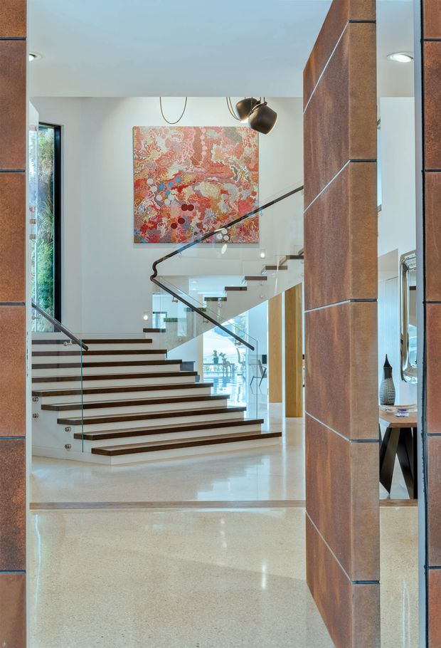 A bright contemporary residential entry and stair hall viewed through a timber-clad opening. A floating staircase with dark timber treads, glass balustrades and slim dark handrails rises toward an upper level. The space has polished light terrazzo or concrete flooring, white walls, tall glazing, timber-lined structural elements, recessed ceiling lights and a sculptural pendant. A large colourful abstract artwork forms a feature on the stair wall, while a dining or living area is visible beyond.