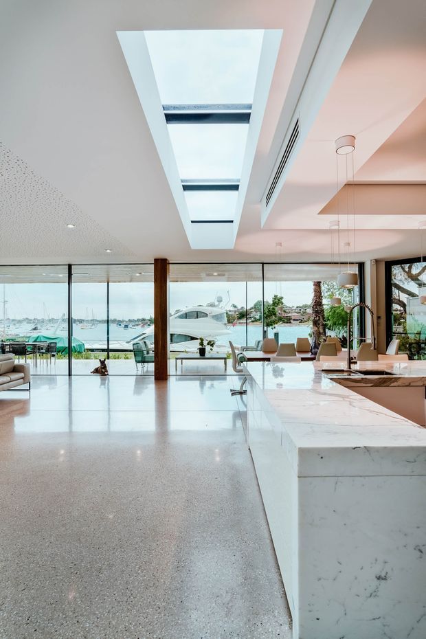 A bright, open-plan waterfront interior combining a modern kitchen, dining area and living space. The room features a large white marble island with waterfall sides, polished terrazzo flooring, extensive floor-to-ceiling glazing, warm timber structural posts, a dramatic linear skylight, and contemporary pendant lighting. Boats and a marina are visible beyond the glazing.