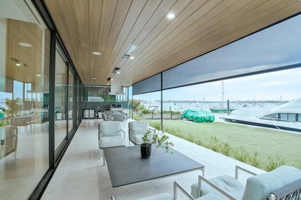 A covered waterfront outdoor living terrace overlooking a marina, with floor-to-ceiling glazing, a timber-lined soffit, pale tiled flooring, contemporary outdoor lounge furniture, and a large dark rectangular coffee table. The space opens directly toward boats and calm water, creating a bright, relaxed coastal setting.