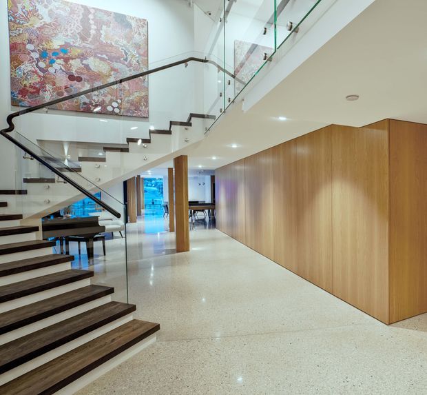 A bright, contemporary residential interior centred on a dramatic open stairway. Dark timber stair treads and a slender dark handrail contrast with clear glass balustrades, white walls and ceiling, and a polished light terrazzo floor. A large colourful artwork forms a feature on the upper stair wall, while extensive warm timber wall panelling and built-in joinery define the right side of the hallway. Dining and living areas are visible beyond the circulation space.