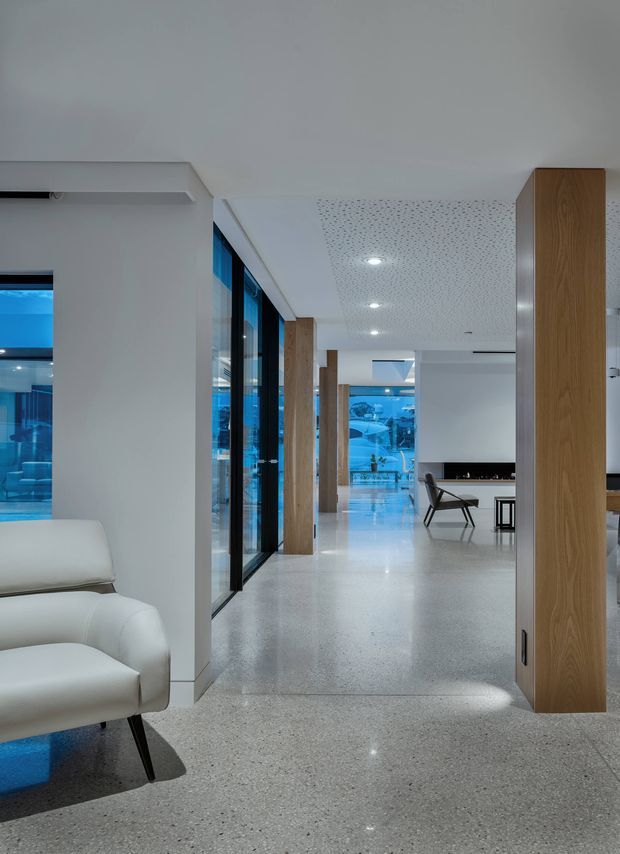 A wide, bright contemporary interior corridor or open-plan living area with polished light terrazzo flooring, white walls, a perforated acoustic ceiling, recessed downlights, and full-height blue-tinted glazing. Warm timber-clad structural or joinery columns line the space, while a white leather lounge chair sits in the foreground and minimalist dining or seating furniture is visible deeper in the room. The composition emphasizes a long open sightline and clean architectural detailing.