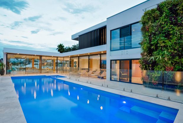 Contemporary two-storey residence viewed from the pool courtyard, featuring crisp white rendered walls, extensive floor-to-ceiling glazing, a projecting upper level with dark vertical screening, and a large illuminated rectangular swimming pool with frameless glass balustrades. A prominent living green wall adds texture to the facade, while warm interior lighting creates an inviting evening atmosphere.