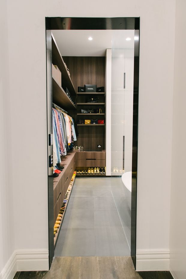 A narrow walk-in wardrobe viewed through a full-height black-framed doorway. The space has dark wood-grain built-in cabinetry, open shelving, drawers, hanging rails filled with clothing, extensive shoe storage, and glossy white tall cupboards. Large grey floor tiles and recessed ceiling lights create a polished, highly organised dressing area. A partial white curved bathroom fixture is visible at the right edge.