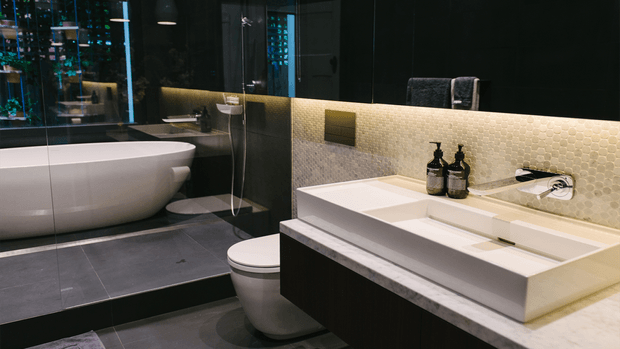 A luxurious contemporary bathroom featuring a freestanding white bathtub behind a glass partition, a wall-mounted white toilet, and a large rectangular vessel basin on dark fixed cabinetry. The space uses dark stone-look wall and floor surfaces, a pale textured mosaic tile backsplash with integrated warm LED lighting, black-framed glazing or mirrors, and chrome fixtures. Reflections reveal additional bathroom surfaces and lighting, creating a moody, refined atmosphere.