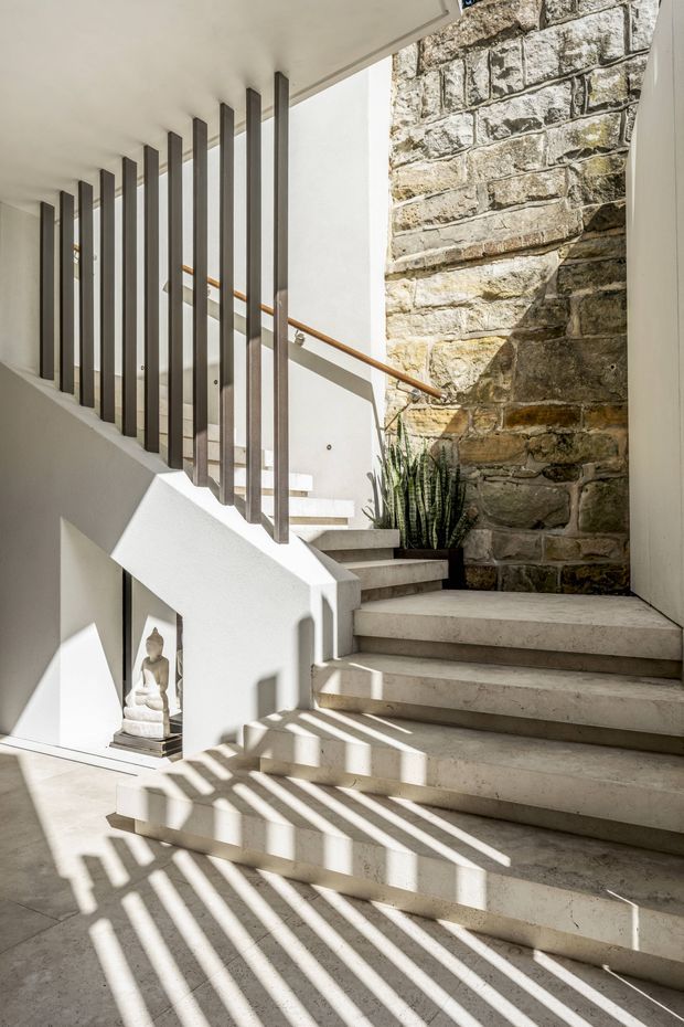 Bright architectural interior photograph of a stairway with broad pale stone or concrete steps, white walls, slim dark vertical metal balusters, and a warm timber handrail. A large exposed rough stone masonry wall forms a strong feature surface beside the stairs, while sunlight casts dramatic linear shadows across the floor and steps. A small sculptural object and potted plant sit near the stair landing.