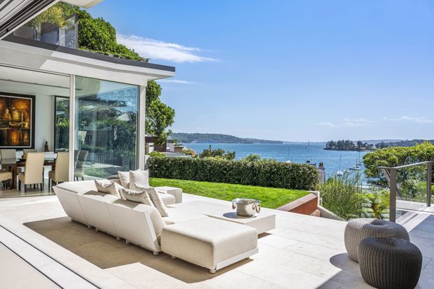 A bright, contemporary waterfront outdoor living terrace overlooking a calm bay. The image shows a spacious paved patio with cream modular lounge seating, an ottoman, and textured dark outdoor chairs. Large sliding glass doors open to an indoor dining area, while layered hedging, lawn, and planted screening frame the view. The house has crisp white rendered walls, strong horizontal rooflines, extensive glazing, and a landscaped elevated site.