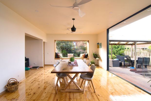 Bright open-plan dining area with a long solid timber dining table and contemporary chairs, polished timber flooring, warm off-white walls, ceiling fans, and large black-framed glazing opening onto a covered timber deck and outdoor dining area. A living space is partially visible to the left and garden greenery is visible beyond the glazing.