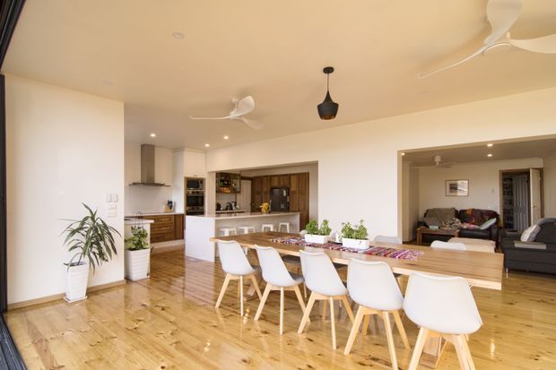 Open-plan contemporary kitchen, dining, and living area with warm timber flooring, white walls and ceiling, a long timber dining table with white molded chairs, and a compact kitchen with timber cabinetry and a peninsula. Recessed lighting and multiple ceiling fans create a bright, practical family interior. A living room with a dark sectional sofa is visible through the wide opening at right.