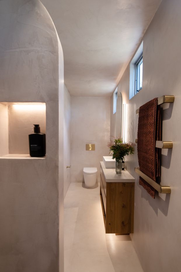 A narrow, refined bathroom with softly curved plastered walls and an arched recessed shelf holding a black soap dispenser. The space features a wall-mounted timber vanity with a white countertop and basin, a concealed toilet, small high windows, warm integrated under-vanity lighting, and brushed brass towel rails holding a textured rust-brown towel.