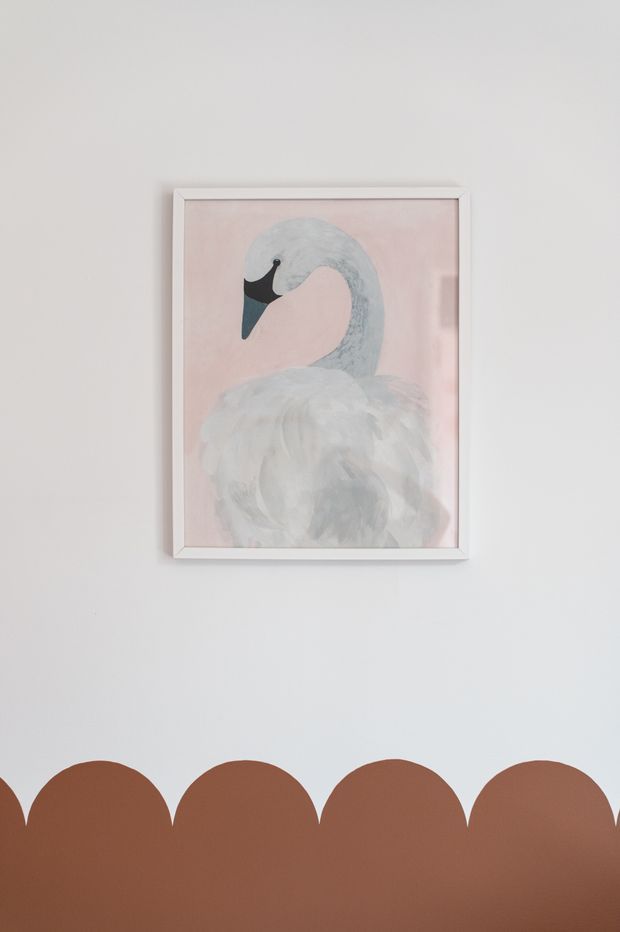 A minimalist interior wall featuring a framed illustration of a swan on a soft pink background. Along the lower wall is a playful scalloped two-tone painted treatment in terracotta brown and pale pink, creating a soft, child-friendly decorative feature.