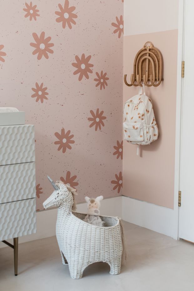 A softly styled children's room corner with blush pink walls, a prominent floral-patterned feature wall, white lower wall panelling, and pale flooring. A woven rattan wall-mounted coat rack holds a small floral backpack, while a white woven animal-shaped basket contains soft toys. A partial white textured cabinet or chest is visible at left. The room has a playful, gentle, warm aesthetic.