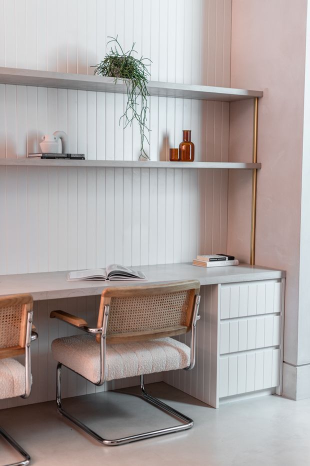 A softly styled built-in study nook with two floating shelves, a long integrated desk, and white vertical panelled wall backing. The desk includes white drawers and cabinetry, with two cantilevered cane-and-upholstered chairs, books, a ceramic vessel, amber glass objects, and a trailing plant. The palette is pale white, blush pink, warm wood, and chrome, with a refined contemporary feel.