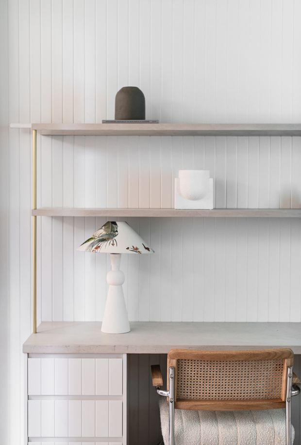 A bright, calm built-in workspace with a white vertical timber-panel wall, pale timber shelving and desktop, integrated drawers, and a cane-and-wood chair. The styling includes a sculptural white table lamp with a botanical-patterned shade and minimal decorative objects.
