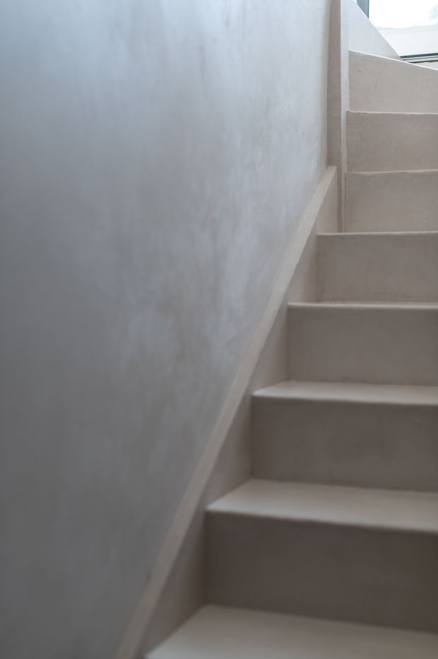 A narrow interior stairway with minimalist plastered walls and fully plastered or concrete-like stair treads and risers. The staircase turns or continues upward toward a bright opening, creating a softly lit, monochromatic architectural scene.
