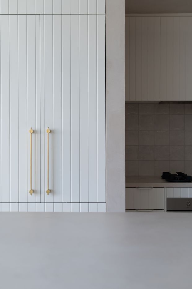 A close, vertically composed view of a restrained contemporary kitchen. White vertical-groove cabinetry with slim brass-gold handles dominates the left side, while a partially visible cooking zone on the right has matching upper and lower cabinetry, a dark grey tiled splashback, and a dark benchtop. A broad pale foreground surface creates a clean, minimalist feel.