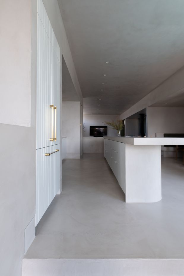 Minimalist open-plan kitchen and living area in a predominantly white and pale grey interior. The space features a long island with a rounded end, integrated white cabinetry with vertical fluting, brass-gold handles, a smooth light floor, recessed ceiling lighting, and a television visible in the background.