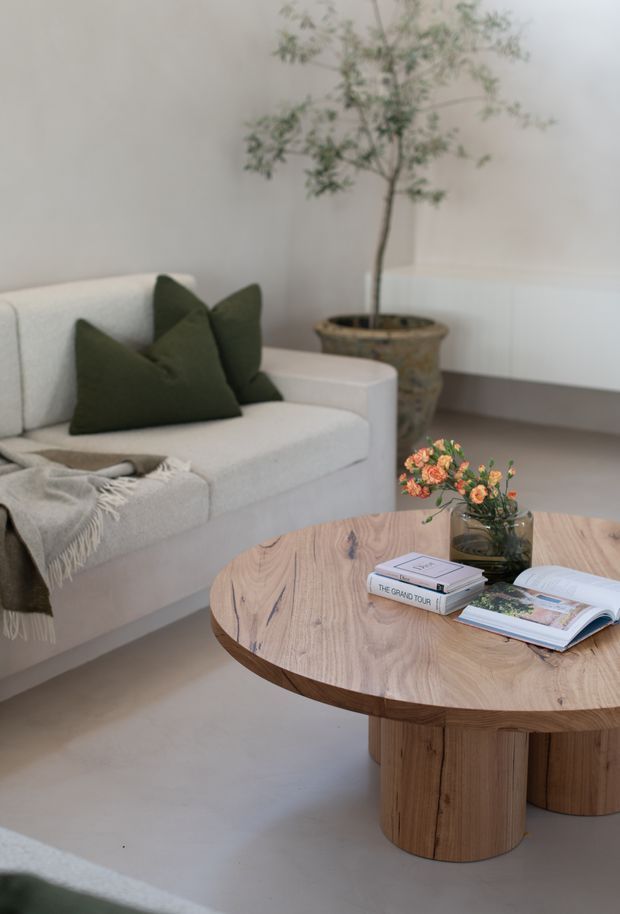 A softly styled living room with a pale upholstered sofa, dark olive-green cushions, a textured throw, and a large rounded timber coffee table with cylindrical pedestal legs. Neutral walls and flooring create a calm, minimalist setting, complemented by a tall potted tree, a small floral arrangement, and books on the table.