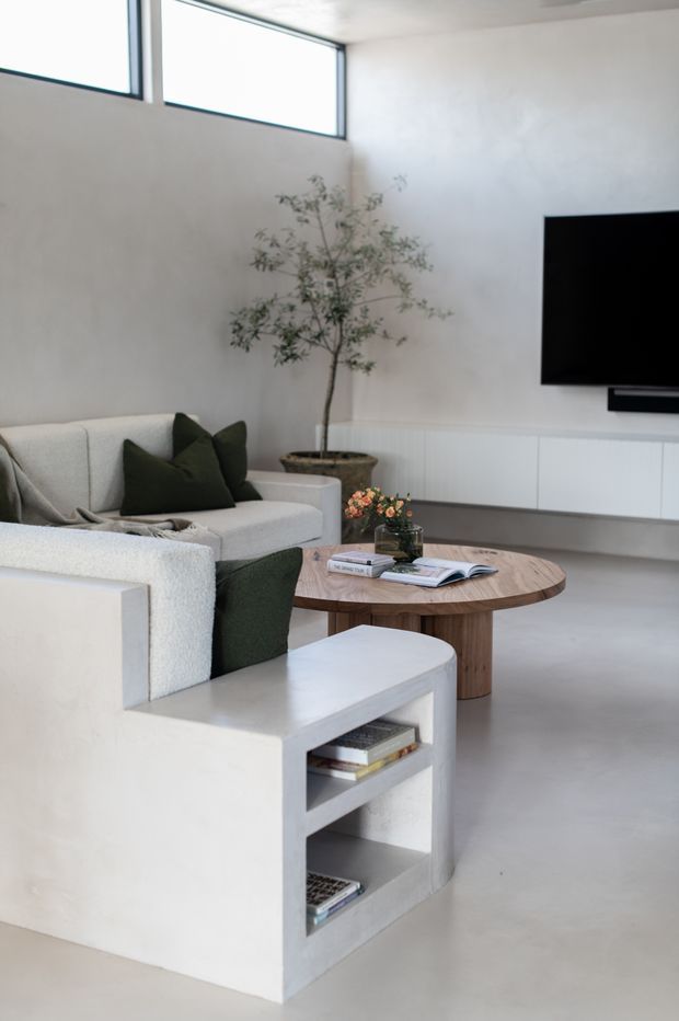 A bright, minimalist living room with a sculptural white built-in lounge chair and open shelving, a light upholstered sofa with dark green cushions, and a round timber coffee table. A wall-mounted television sits above streamlined white cabinetry, with a tall indoor tree adding a natural element. High horizontal windows and smooth pale surfaces create an airy, contemporary interior.