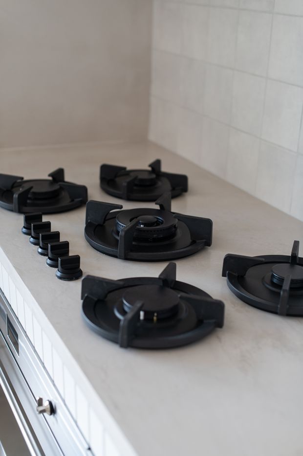 Close-up view of a light kitchen benchtop fitted with multiple black gas burners and matching control knobs. White tiled splashback and white cabinetry are visible, creating a clean, understated kitchen setting.