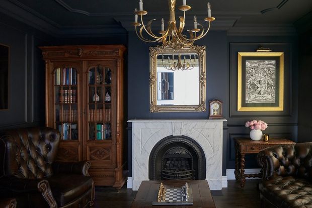A richly styled traditional living room or study with deep navy walls, ornate ceiling mouldings, a white carved fireplace with dark arched surround, an antique gold chandelier, gilt-framed mirror, dark carved timber bookcase, and tufted brown leather seating. The room has a moody, refined library-like atmosphere with classic decorative detailing.