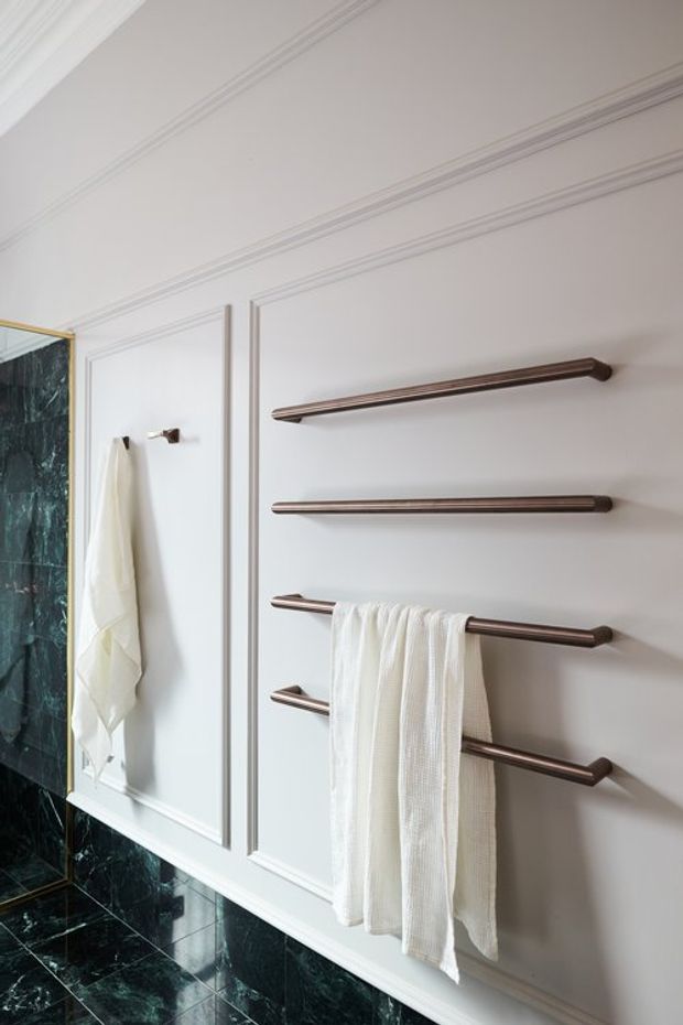 Close-up view of a refined bathroom wall featuring white painted decorative panel moulding, multiple horizontal dark bronze towel rails, a hanging white towel, and glossy dark green marble wall and floor tiles. A partial glass shower enclosure is visible at the left.