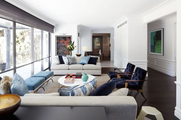 Bright, refined living room with large floor-to-ceiling windows, layered cream and navy seating, a round coffee table, dark timber flooring, white panelled walls and substantial decorative cornices. A vertical timber screen and artwork add visual interest at the rear of the room.