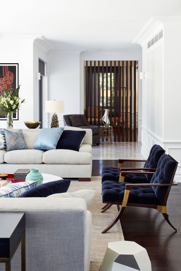 Bright, refined living room with a layered seating arrangement, including a large pale upholstered sofa, navy velvet lounge chairs with timber arms, and patterned cushions. The room features white paneled walls and ceiling cornices, dark timber flooring, a pale textured rug, polished stone flooring beyond the seating area, and a dramatic vertical timber screen dividing the living space from the rear area. Contemporary artwork, greenery, sculptural decor, and soft natural light complete the sophisticated interior.