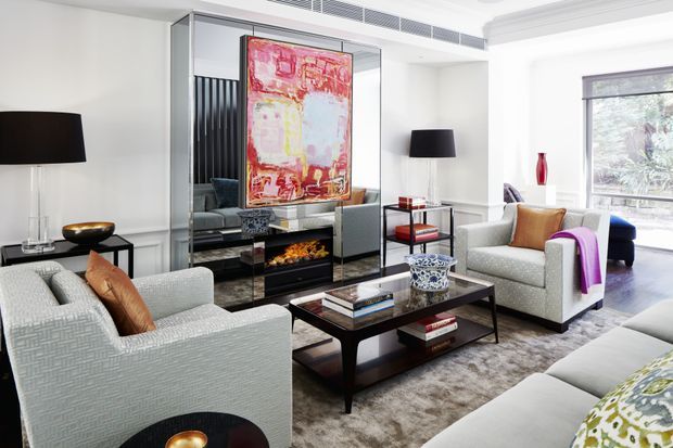 Bright, refined living room with two textured light-grey armchairs, a large sectional sofa, dark wood and glass coffee table, black lamps, a mirrored built-in media wall with an electric fireplace, and a colourful abstract artwork. White walls, substantial layered cornice detailing, and large windows create an elegant contemporary setting.