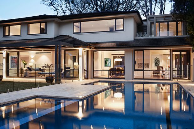 A contemporary two-storey residence viewed from the landscaped pool area at dusk. The home features white rendered walls, dark low-pitched roof planes, extensive black-framed glazing, covered outdoor living areas, and a large rectangular swimming pool with reflective water. Warm interior lighting reveals living, dining, and circulation spaces behind the glass.