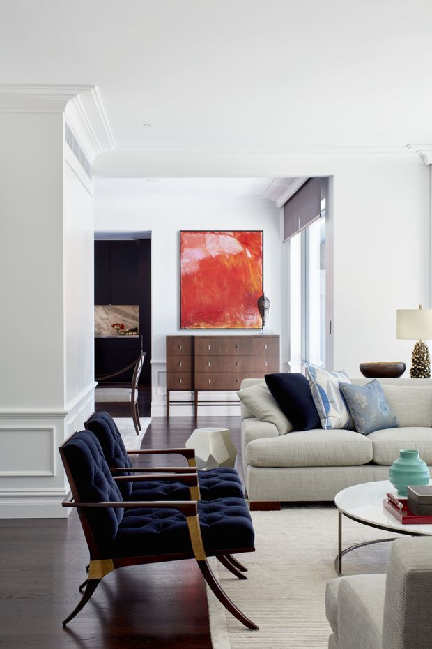 Bright, refined living room with white walls, ornate ceiling cornices and wall mouldings, dark timber flooring, and large windows with roller blinds. A light upholstered sofa and pair of deep navy lounge chairs surround a round coffee table, while a timber sideboard and bold red-orange artwork form a focal point in the adjoining area. A dark kitchen or dining space is visible through the opening.