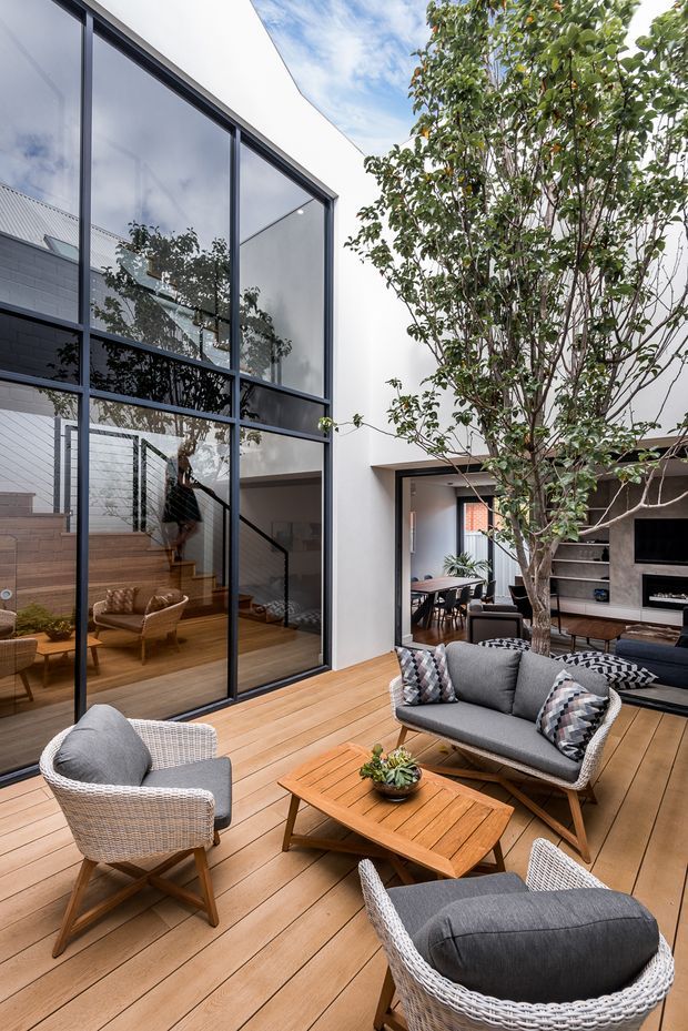A contemporary courtyard-style outdoor living area with a timber deck, woven outdoor lounge chairs, a grey sofa, and a timber coffee table. Floor-to-ceiling black-framed glazing opens the deck to a bright modern interior with a visible stairway and dining area. A mature tree occupies the central courtyard, while white rendered walls and dark brick interior surfaces create a clean, architectural backdrop.
