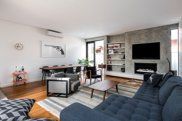 A bright, contemporary open-plan living and dining area with white walls, warm timber flooring, and a textured grey feature wall incorporating built-in shelving, a wall-mounted television, and a linear fireplace. A large dark blue sectional sofa, patterned area rug, timber coffee table, dining table with dark chairs, armchairs, indoor plants, and restrained decorative furnishings create a refined yet comfortable interior.
