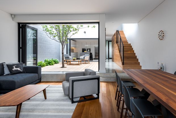 A refined open-plan living and dining area with a dark upholstered sofa, sculptural armchairs, timber coffee table, and a long solid-wood dining table with black chairs. Large full-height glazed openings connect the interior to a central landscaped courtyard and a secondary seating area. A floating timber staircase with black balustrade rises along a white wall, while warm timber flooring and restrained contemporary detailing create a bright, spacious atmosphere.